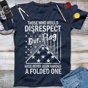 May include: A navy blue t-shirt with a distressed white graphic of an American flag folded in a triangle. The text "Those who would disrespect our flag have never been handed a folded one" is printed below the flag.
