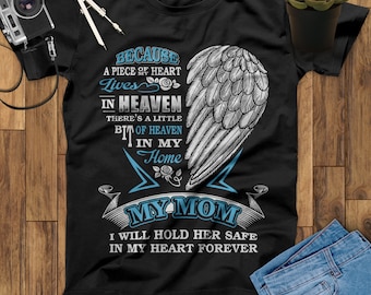 Op de afbeelding: Zwart T-shirt met een wit engelenvleugelontwerp en de tekst "Omdat een deel van mijn hart in de hemel leeft, is er een beetje hemel in mijn huis, Mijn Moeder, ik zal haar voor altijd veilig in mijn hart houden".