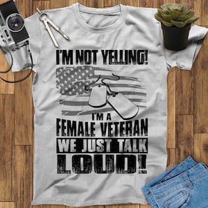 May include: Gray t-shirt with a black and white graphic design. The graphic features a stylized American flag, two dog tags, and the text "I'm not yelling! I'm a female veteran we just talk loud!"