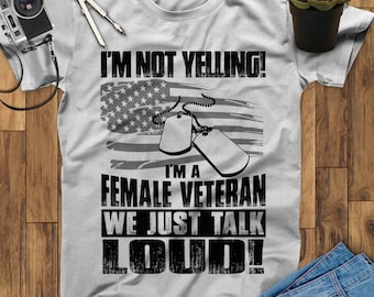 May include: Gray t-shirt with a black and white graphic design. The graphic features a stylized American flag, two dog tags, and the text "I'm not yelling! I'm a female veteran we just talk loud!"