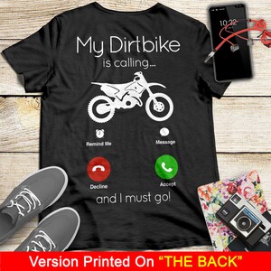 May include: Black t-shirt with a white graphic of a dirt bike with the text "My Dirtbike is calling..." and a phone icon with the options "Remind Me", "Message", "Decline", and "Accept". The text "and I must go!" is below the phone icon.