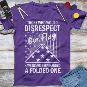 May include: Purple t-shirt with a white distressed graphic that says "Those who would disrespect our flag have never been handed a folded one". The graphic includes a folded American flag with stars.