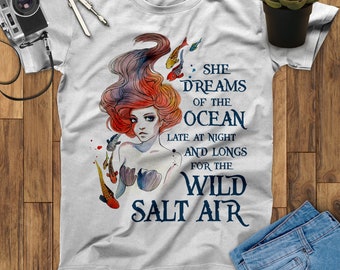 May include: A white t-shirt with a watercolor illustration of a mermaid with red hair and a tail. The text "She dreams of the ocean late at night and longs for the wild salt air" is printed on the shirt.