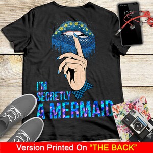 Peut inclure: T-shirt noir avec un motif d'&eacute;cailles de sir&egrave;ne bleu et violet et le texte "I'm Secretly A Mermaid". Un doigt avec un ongle bleu est montr&eacute; avec son index sur ses l&egrave;vres, comme pour dire "chut".
