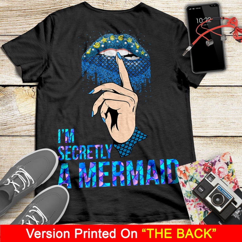 Peut inclure: T-shirt noir avec un motif d'&eacute;cailles de sir&egrave;ne bleu et violet et le texte "I'm Secretly A Mermaid". Un doigt avec un ongle bleu est montr&eacute; avec son index sur ses l&egrave;vres, comme pour dire "chut".