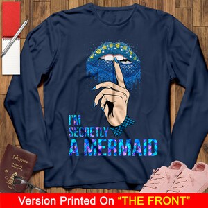 Peut inclure: Un t-shirt &agrave; manches longues bleu marine avec un motif sur le th&egrave;me de la sir&egrave;ne. Le motif repr&eacute;sente une l&egrave;vre de sir&egrave;ne bleue et violette avec un doigt dessus, et le texte "I'm Secretly A Mermaid".