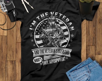 Puede incluir: Camiseta negra con un gráfico blanco de un soldado sosteniendo un rifle frente a una bandera estadounidense. El texto dice "Soy el veterano y el esposo del veterano. ¿Cuál es tu superpoder?"