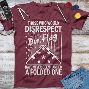 May include: A maroon t-shirt with a white distressed graphic that reads "Those who would disrespect our flag have never been handed a folded one". The graphic includes a folded American flag with stars and stripes.