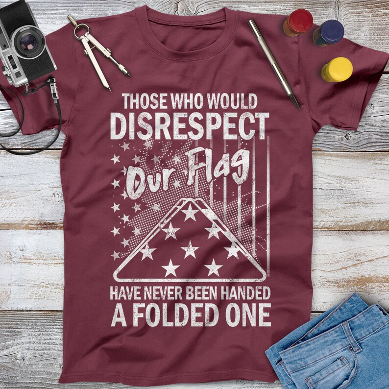 May include: A maroon t-shirt with a white distressed graphic that reads "Those who would disrespect our flag have never been handed a folded one". The graphic includes a folded American flag with stars and stripes.