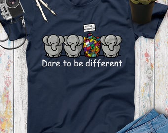 May include: A navy blue t-shirt with a graphic of three elephants. The middle elephant is holding a sign that says "Autism Awareness" and is made up of puzzle pieces. The text "Dare to be different" is printed below the elephants.