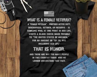 Puede incluir: Camiseta negra con un estampado gráfico blanco que dice "What is a female veteran?" e incluye una definición del término. El gráfico también incluye una pequeña bandera estadounidense. El texto en la parte inferior del gráfico dice "That is honor."