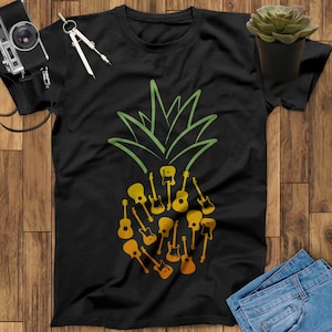 May include: Black t-shirt featuring a green pineapple top design with a body made of yellow guitars. The guitars are in various shapes and sizes. The shirt is displayed on a wooden surface with a camera, compass, succulent, and jeans.