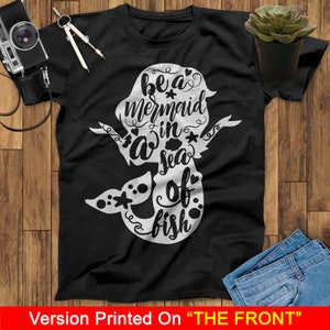 以下が含まれることがあります： 黒いTシャツに、白いマーメイドのグラフィックと「Be a mermaid in a sea of fish」というテキストがプリントされています。