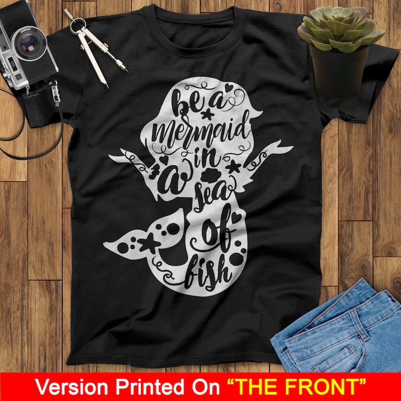以下が含まれることがあります： 黒いTシャツに、白いマーメイドのグラフィックと「Be a mermaid in a sea of fish」というテキストがプリントされています。