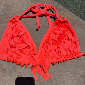 May include: A bright neon orange bikini top featuring a classic triangle silhouette with thin halter neck ties. The front panels are adorned with numerous vertical fabric fringe strips that create a textured, layered appearance across the chest.