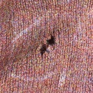 May include: Close-up of a textured, reddish-brown knit fabric with a hole. The fabric has a mix of colors, including red, brown, and hints of yellow and blue. The knit pattern is visible, and the hole reveals the inner structure of the fabric.