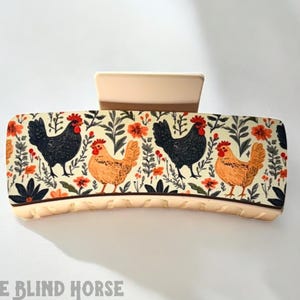 May include: A cream-colored hair claw with a floral pattern featuring black and orange chickens. The claw has a textured surface and a small, cream-colored bar at the top.