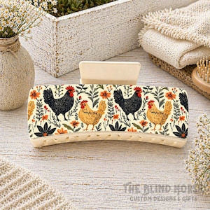 May include: A cream-colored hair claw clip featuring a repeating pattern of black and brown chickens, orange flowers, and green leaves. The clip has a curved shape with a smooth, matte finish, and is suitable for various hair types.