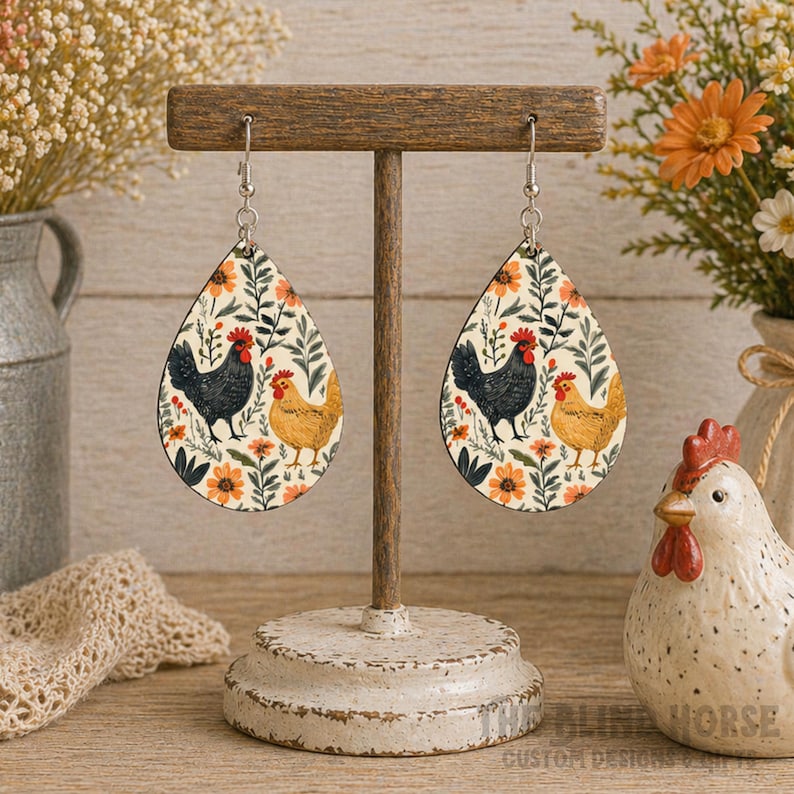 K&ouml;nnte beinhalten: Tropfenf&ouml;rmige Ohrringe mit cremefarbenem Hintergrund, die ein schwarzes und ein braunes Huhn zeigen, umgeben von orangefarbenen Blumen und gr&uuml;nen Bl&auml;ttern. Die Ohrringe werden auf einem Holzst&auml;nder pr&auml;sentiert, der einen rustikalen, l&auml;ndlichen Stil widerspiegelt.