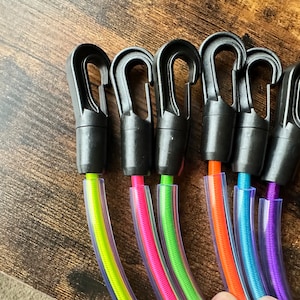 May include: Six black plastic swivel snap hooks attached to colorful bungee cords encased in clear plastic tubing. The cords are neon yellow, hot pink, lime green, bright orange, sky blue, and purple, each featuring a spring-loaded gate mechanism.