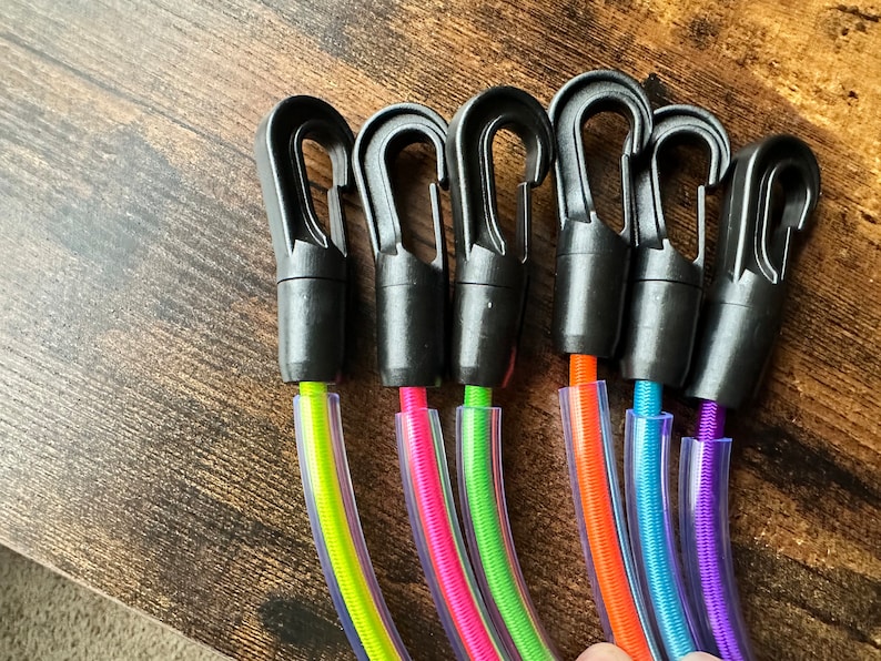 May include: Six black plastic swivel snap hooks attached to colorful bungee cords encased in clear plastic tubing. The cords are neon yellow, hot pink, lime green, bright orange, sky blue, and purple, each featuring a spring-loaded gate mechanism.