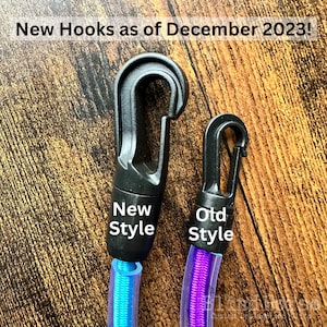 May include: Two black plastic swivel hooks attached to colorful cords. The larger hook is labeled New Style and the smaller hook is labeled Old Style. Text at the top reads New Hooks as of December 2023! The cords are blue and purple.
