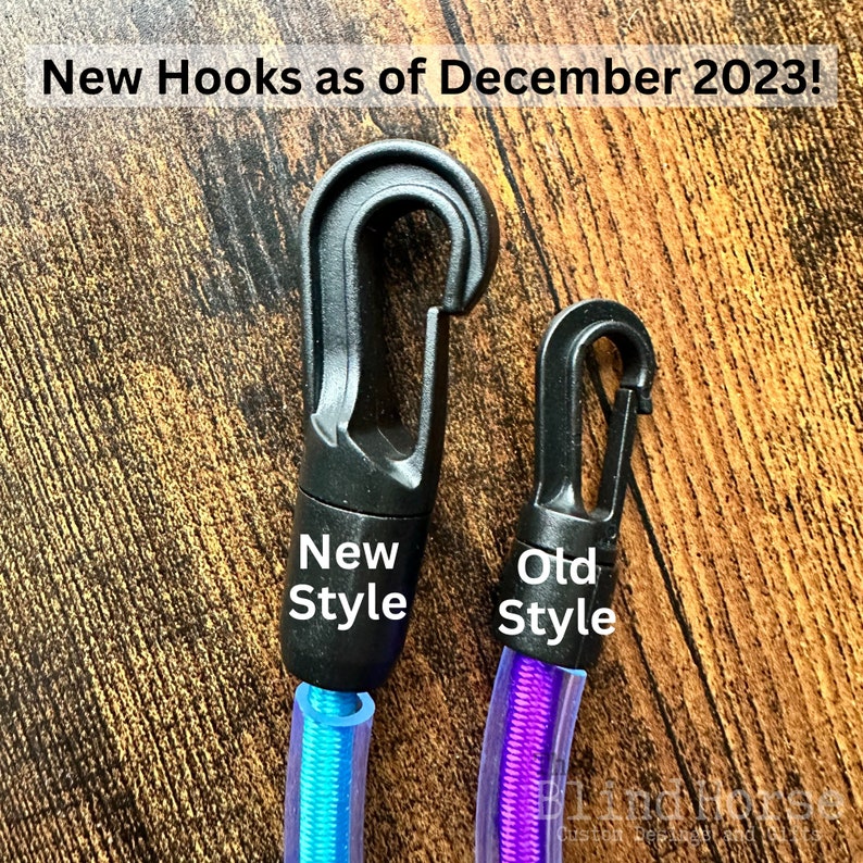 May include: Two black plastic swivel hooks attached to colorful cords. The larger hook is labeled New Style and the smaller hook is labeled Old Style. Text at the top reads New Hooks as of December 2023! The cords are blue and purple.