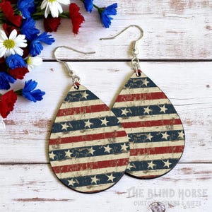 Puede incluir: Pendientes en forma de l&aacute;grima con un dise&ntilde;o patri&oacute;tico. Los pendientes presentan un patr&oacute;n desgastado de rayas rojas y azules con estrellas blancas, evocando la bandera americana. Los pendientes tienen ganchos plateados.