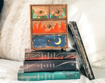 May include: A stack of four paperback books with the title "A Court of Thorns and Roses" and the author's name "MAAS" on the spines. The books are stacked on top of a small wooden dresser with three drawers. The drawers are painted with a floral design, flames, and a night sky with a crescent moon.