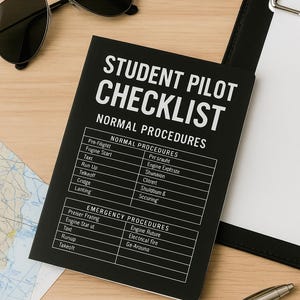 May include: A black student pilot checklist notebook with white text, including normal and emergency procedures. The notebook is on a wooden surface with sunglasses, a pen, a map, and a clipboard.