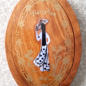 Op de afbeelding: Een ovale houten sieradendoos met een scharnierend deksel. Het deksel heeft een geschilderd beeld van een vrouw in een traditionele Vietnamese jurk, met de woorden "Viet Nam" in goud erboven geschreven. De doos is versierd met een gouden rand en bloemmotieven.