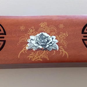 Peut inclure: Une boîte rectangulaire en bois avec une finition brun rougeâtre. La boîte présente un motif floral central avec une rose blanche et des accents dorés. Deux motifs noirs découpés en cercle se trouvent à chaque extrémité de la boîte.