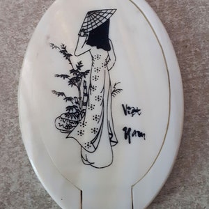 Pode incluir: Espelho compacto ovalado branco sujo com uma ilustração a preto e branco de uma mulher em traje tradicional vietnamita, usando um chapéu cónico. O texto "Viet Nam" está escrito a preto.