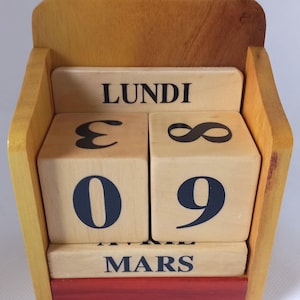Op de afbeelding: Houten kalender met de aanduiding "LUNDI" en "MARS". De datum wordt weergegeven als "09" op houten blokken. De kalender is gemaakt van lichtbruin hout met een rode basis.