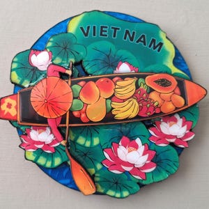Pode incluir: Um íman de lembrança do Vietname colorido. O íman apresenta um barco cheio de frutas tropicais, uma pessoa com um guarda-chuva laranja e flores de lótus. A palavra "VIETNAM" está impressa em verde no topo.