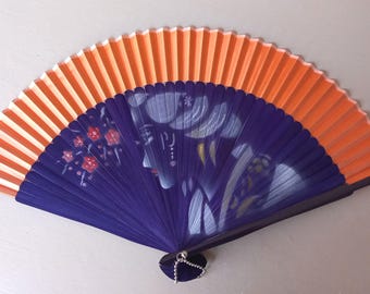 May include: A folding hand fan featuring a vibrant orange upper section and a deep purple lower section. The purple area displays a stylized illustration of a person in profile with white hair details and small red floral accents near the left side.