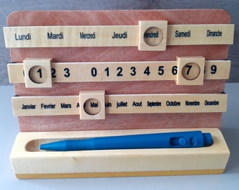 May include: A wooden calendar with a pen holder. The calendar has days of the week, months of the year, and numbers 1 through 9. The pen holder is made of light wood and has a blue pen inside.