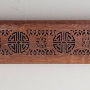May include: A wooden incense holder with intricate carvings of geometric patterns and a repeating symbol. The holder is brown and has a rectangular shape.