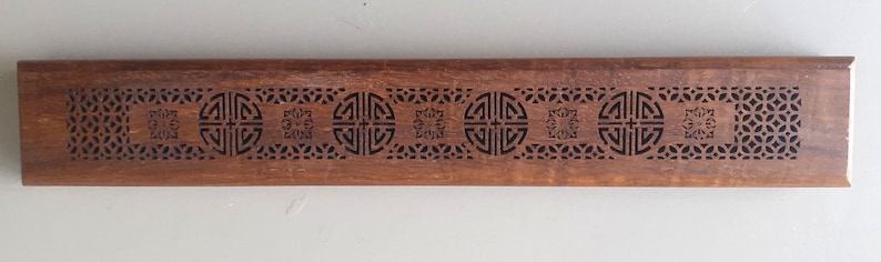 May include: A wooden incense holder with intricate carvings of geometric patterns and a repeating symbol. The holder is brown and has a rectangular shape.