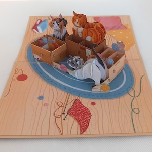 Puede incluir: Una tarjeta de felicitación desplegable con una escena tridimensional de gatos jugando con lana en cajas de cartón. La ilustración incluye un gato calicó, un atigrado naranja y un gato gris sobre una alfombra azul, con agujas de tejer y lana roja.