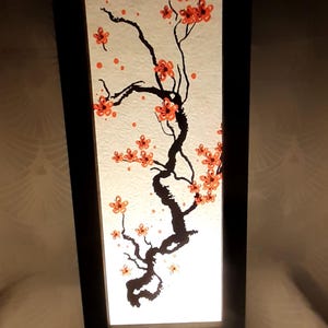 以下が含まれることがあります： オレンジの花が咲いた黒いシルエットの桜の木が描かれた白い紙のシェードが付いた黒い木製のテーブルランプです。