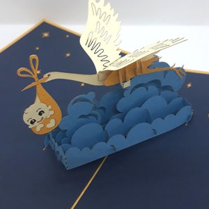 Op de afbeelding: Een driedimensionale pop-up wenskaart met een crèmekleurige ooievaar die boven gelaagde blauwe wolken vliegt. De ooievaar draagt een klein, oranje bundeltje met een baby. De basis is een donkerblauwe kaart met gouden sterren.