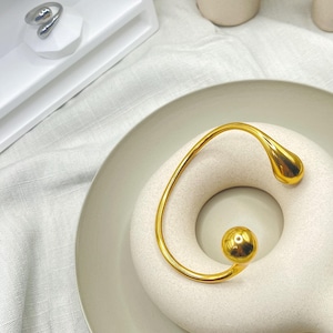 May include: A gold-toned abstract ring with a rounded, teardrop-shaped design. The ring is displayed on a white ceramic base.