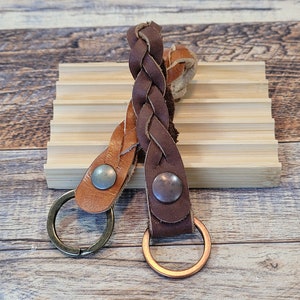 Op de afbeelding: Twee leren sleutelhangers met gevlochten leren bandjes. De ene sleutelhanger is bruin met een bronzen ring, de andere is bruin met een zilveren ring. Beide sleutelhangers hebben een metalen drukknoopsluiting.