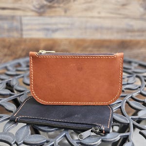 May include: Two leather coin purses. The top purse is a light brown with a gold zipper and white stitching. The bottom purse is black with a silver zipper. Both purses are rectangular with rounded corners.