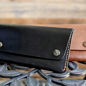 May include: Two leather wallets, one black and one brown, are displayed. The black wallet is in the foreground, with two silver snap closures. The brown wallet is behind it, with a single snap closure. Both wallets are rectangular and appear to be handmade.