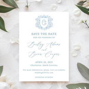 May include: A white wedding invitation with blue text and floral accents. The text reads "Save the Date" and lists the names Bailey Adams and Brian Cooper, along with the wedding date and location. The invitation is surrounded by white petals and green leaves.