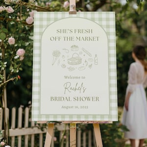 May include: A sign on an easel with a green and white gingham border. The sign reads "She's Fresh Off The Market" and "Welcome to Rachel's Bridal Shower August 16, 2025." The sign has illustrations of picnic items.