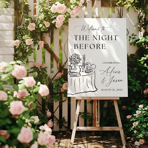 May include: A white sign on a wooden easel with the text "Welcome to The Night Before" and the names Alexis & Jason, celebrating August 21, 2025. The sign is surrounded by pink roses and greenery.