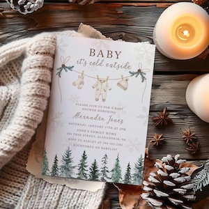 May include: A baby shower invitation with the text "BABY it's cold outside." The invitation features illustrations of baby clothes hanging on a clothesline, snowflakes, and a forest scene. The invitation is surrounded by candles and pinecones.